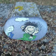 Un petit personnage rêveur s’est posé près d’une grosse pierre grise. Cherche au bord du gravier, là où la mousse colore les blocs… son grand sourire n’attend plus que toi !
